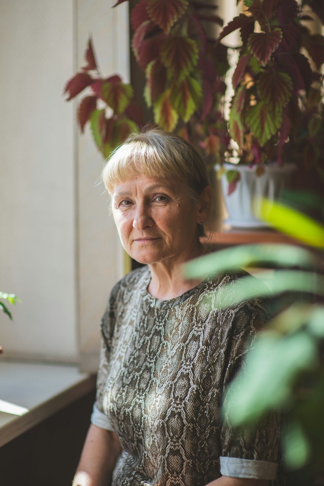 Een vrouw met grijs haar staat bij een raam met planten